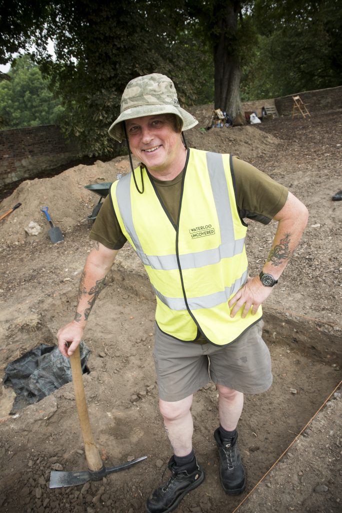 Jim Howdle on site in 2019, holding a mattock and standing in a trench