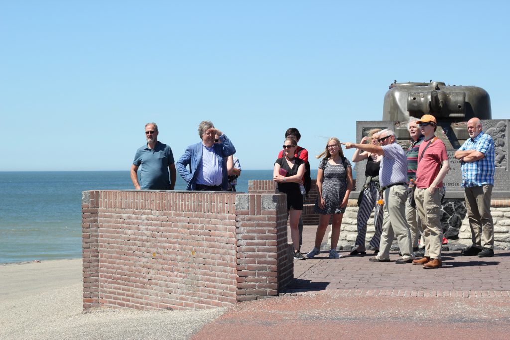A group of summer school participants on a field trip, standing in front of a war memorial on the coast