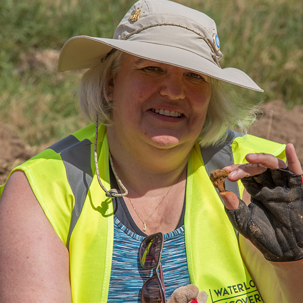 Emma Wray Smith in 2019 at Mont Saint Jean, holding up a find she discovered, wearing a Hat with Navy pins and a yellow high vis jacket.